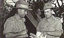 Two men in military uniform conferring in front of a tent. The one on the right is holding a stack of papers.