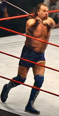 A white man running, wearing blue shorts and boots, while in a professional wrestling four-sided ring holding a pole with both hands.