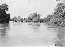 7 soldiers standing on pontoon bridge in middle distance; fast flowing wide River Jordan in foreground; lush growth on both banks.