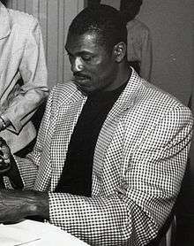 A man, wearing a checkered coat and a dark shirt, is signing an autograph.