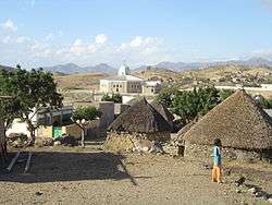 A small village in a dry environment. There are some round stone huts in the foreground with conical thatched roofs. In the middle distance is a modern octagonal church with a white dome and cross. There are a few trees scattered in the village, and a donkey and child in the foreground.