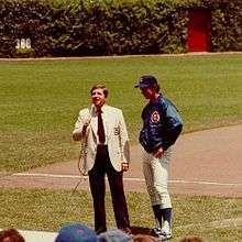 Two men standing on a grass-and-dirt field; the man on the right is wearing a blazer and slacks, while the man on the left&mdash;the subject of the image&mdash;is wearing white pinstriped baseball pants, a blue nylon warm-up jacket, and a blue baseball cap.
