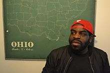 Hanif Willis-Abdurraqib pictured in front of a map of Ohio. He wears a black shirt and jacket and a red baseball cap, turned backwards, and looks off-camera.
