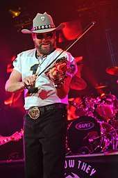 A bearded man wearing a cowboy hat and dark glasses, playing a violin