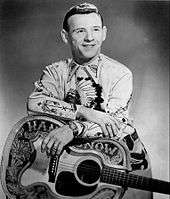 A man wearing an elaborately patterned shirt, leaning on a guitar