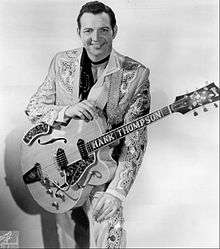 A dark-haired man wearing an elaborately-patterned suit and holding a guitar