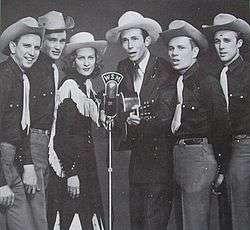 A group of six men and one woman wearing cowboy hats, standing around a microphone.  The man third from right is holding a guitar.