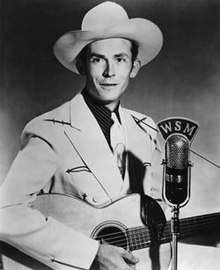 A man in a light jacket and cowboy hat, playing a guitar at a microphone