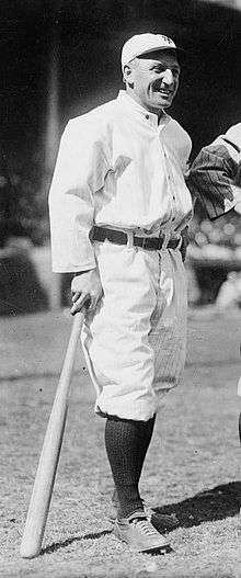 A smiling man wearing an old-style white baseball uniform and cap, standing on a baseball field and leaning on a baseball bat
