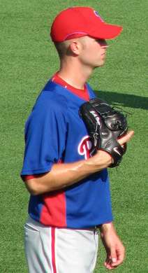 A young man in a blue baseball jersey and red baseball cap holds a black baseball glove against his chest with his left hand