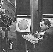 A young man sits holding a microphone in his left hand while manipulating the console of an apparatus with his right. To his left a large television camera is trained on a large, circular cathode ray tube display.