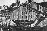 Two large wooden buildings, each about 60 to 80 feet across, are being constructed into a hillside. About five people stand on the roof of the nearest building while below, upwards of 15 people stand amidst piles of long timber. The exteriors of the buildings appear to be almost complete, with only the lower third of the nearest building still showing exposed framing.