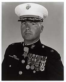 Head and shoulders of a middle-aged white man wearing a white peaked cap with oak leaf decorations on the visor and a dark military jacket with two rows of medals hanging from ribbons pinned to his chest and another medal hanging from his neck.