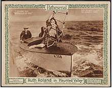 Kneeling on the prow of a speeding boat, Ruth struggles to climb a ladder suspended from an airplane.