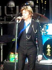 A male singer wearing a dark grey three-piece suit, white shirt and a blue undone tie, on stage singing while holding a microphone on a stand.
