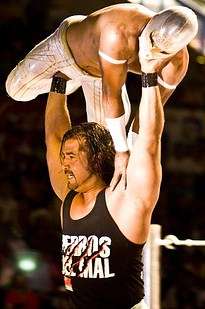 An unmasked wrestler lifting a masked wrestler up over his head with outstretched arms.