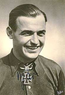 The head and shoulders of a young man, shown in semi-profile. He wears a shirt with an Iron Cross displayed at the front of his shirt collar.