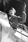 A pilot seated in his fighter craft gestures with a gloved hand.