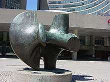 Photograph of a large bronze abstract sculpture, in front of a glass and concrete building.