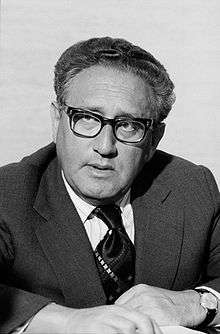 A head-and-shoulders photograph, in grayscale, of Henry Kissinger, looking to the viewer's left with a thoughtful expression on his face.