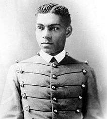 Cadet Henry O. Flipper in his West Point cadet uniform. It has three large round brass buttons left, middle and right showing five rows. The buttons are interconnected left to right and vice versa by decorative thread. He is wearing a starched white collar and no tie. He is a lighter colored African-American with plaited corn rows of neatly done hair. He is facing the camera and looking to the left of the viewer.