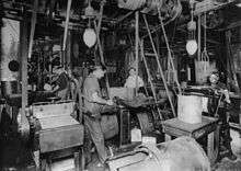 Photograph of a large workshop crowded with machinery. There are at least four men working there. There are belts coming down from the ceiling that drive the machinery. Two electric lighting fixtures are also hung from the ceiling.