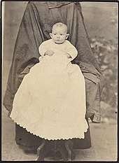 Baby in a long white dress on a chair draped plain dark fabric that may be two coats.