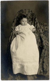 Baby in a long white dresss at on what appears to be a chair draped in fabric.