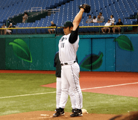 A Japanese man wearing a Devil Ray baseball uniform points his arms upward as he prepares to pitch in the bullpen.