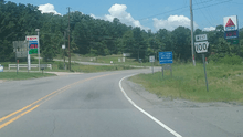 Roadway in a rural area with two gas station signs and trees in the background, and a "Arkansas Highway 100" sign in the foreground.