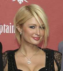A Caucasian woman seen from bust level up with short blonde hair wearing a lacey, strappy black top with a silver necklace, looking slightly askance at the camera and smiling faintly, in front of a reddish-pink background with some fragments of words on it
