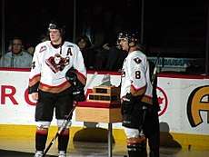 Two players in full hockey uniforms stand with a trophy between them.