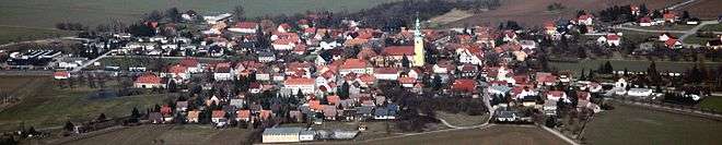 A 180-degree view of a large village surrounded by gently sloping fields. A church steeple rises from the center of the village.