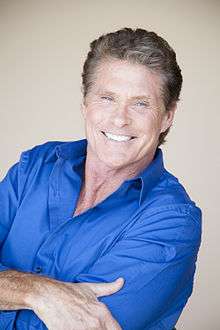 An image of Hasselhoff in a blue collared shirt smiling straight to a camera with his arms crossed