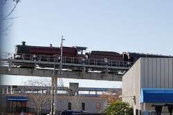 The lower half of the photograph shows industrial office buildings, with a small label "T-4" in one corner.  Above this runs a steel construction carrying an elevated rail system, with an emergency exit walkway.  Mounted on the track is a red steam locomotive with two small wheels and three big wheels raised slightly above the track.  Above the middle of the three big wheels is a curved sign that says "Hogwarts Castle".  Under the train are sets of much smaller wheels resting directly on the track.