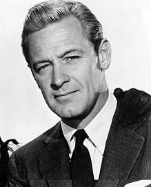 William Holden in a publicity photo in 1954—a white man with light hair and small eyes, with a faint smile, wearing a suit, around 40 years of age.