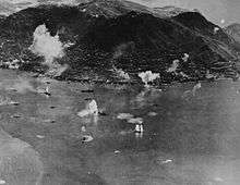 Aerial photo of an island with an urban area along its shore and a steep mountain in the center. A large number of ships are in the water next to the island, and plumes of water are erupting near some of them.
