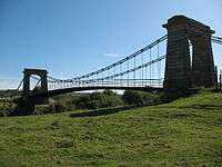 Suspension bridge with stonework arches either end, partly silhouetted against clear blue sky, green field in foreground