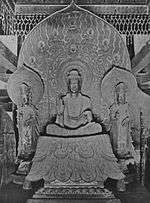 Front view of a central figure sitting cross-legged on a raised platform which is flanked by two smaller standing statues. The central figure has the palm of his right hand turned to the front. The attendants look identical pointing upwards with their right hand and their left hand lifted halfway touching the thumb with the middle finger. Each of the three statues has a halo.