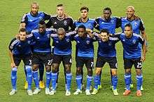 Eleven soccer players wearing blue jerseys and black shorts standing in two lines posing for a pre-match photo.