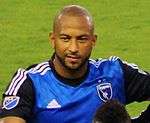 A man with a shaven head wearing a blue soccer jersey.
