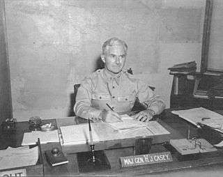 A name plaque reads: "Maj. Gen. Hugh J. Casey". The wall behind him is covered in huge map of New Guinea.