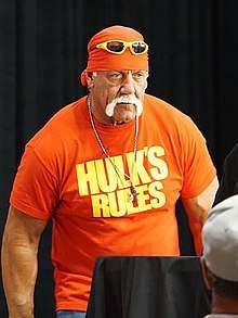 A muscular Caucasian man in a red T-shirt with "Hulk's Rules" written on it in large yellow letters, a red bandanna with yellow-trimmed sunglasses pulled up on it and a lengthy white handlebar mustache stands in front of a black curtain, looking slightly downward.
