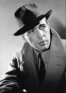 Black-and-white photo of Humphrey Bogart in 1940—a charming white man with dark eyes and a square face, wearing a dark hat and a light-colored overcoat, around 35 years of age.