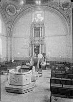 A sepia photograph shows the interior of the synagogue. In the foreground lies the reading desk atop a simply-paneled almemar. Rows of wooden benches line the right and left side of the nave. The holy ark, set in the centre of the eastern wall, is decorated with baroque carvings and set off against four Corinthian columns. Its top reaches a large clover-shaped window, which sits just below one of the four supporting arches. The walls faintly show decorative murals, with two large round frescos situated at the top left and right corners.