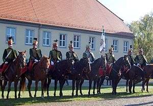 Photo of eight horsemen dressed in Austrian hussar uniforms of the early 1800s, with red shako, dark green jackets, and red breeches