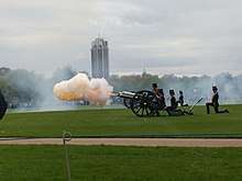 A cannon of the King's Troop Royal Horse Artillery firing in Hyde Park. A large plume of orange smoke, lit orange by the flash from the gun, extends from the barrel.