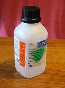 White plastic bottle With safety cap, labeled "QP Panreac" above smaller text "Hydrofluoric Acid 40% QP" with 6 translations. In a bright orange region along the side, warning symbols are visible.