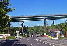 A pair of bridges pass from left to right on tall supports that tower over the other structures in the area, such as telephone poles and single-story buildings. The two-lane NY&nbsp;22 passes underneath the bridge and proceeds into the background.