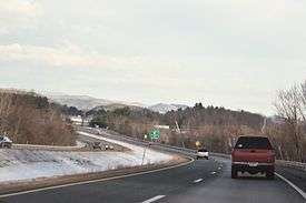 A four lane highway in snowy weather curving left with several cars on it. An exit sign and mountains are in the distance.